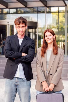 business man and woman standing outside of an office building smiling happy while looking at camera, they are wearing casual clothes and she has a briefcase
