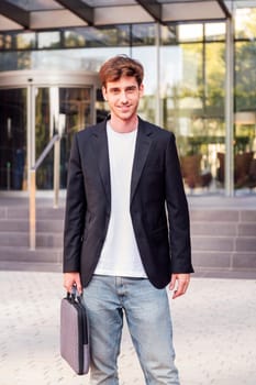 young adult man in casual suit standing looking at camera holding a briefcase in front of an office building, concept of entrepreneur and business
