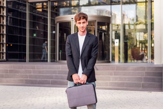 young adult man in casual suit standing looking at camera holding a briefcase in front of an office building, concept of entrepreneur and business, copyspace for text