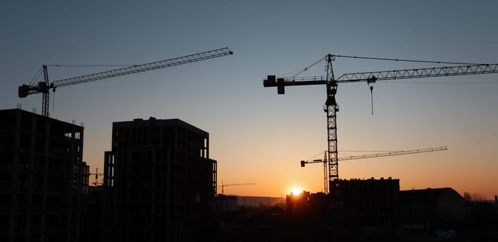 Construction cranes tower over a developing urban area as the sun sets, casting a golden glow on the buildings. The cranes are actively working, signifying ongoing projects.