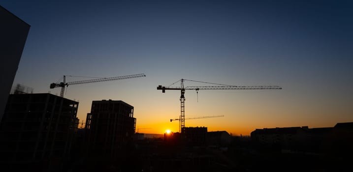 A vivid sunset casts warm hues over a construction site, where cranes stand tall against the skyline. The silhouettes of buildings create a dramatic contrast in the fading light.