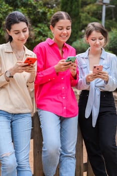 Three young Caucasian women on a wooden bench in a green park,smiling while using smartphones.they look happy and engaged,enjoying a casual outdoor moment with modern devices and social media apps