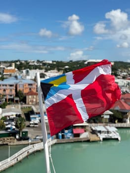Vibrant nautical flag flutters in the breeze above a tropical Caribbean harbor, with pastel buildings, turquoise water, and a sunny blue sky in the background.