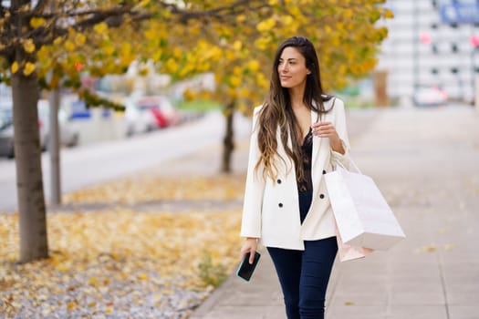 A fashionable woman strolls along a busy sidewalk, holding colorful shopping bags, surrounded by autumn foliage, showcasing her elegance and leisurely lifestyle with a joyful demeanor