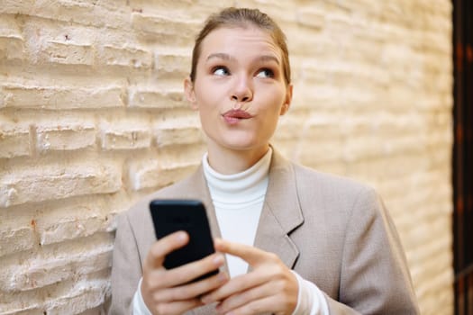 A youthful woman, wearing a contemplative and pensive look on her face, is absorbed in using her smartphone while casually leaning against a rugged and textured brick wall in broad daylight