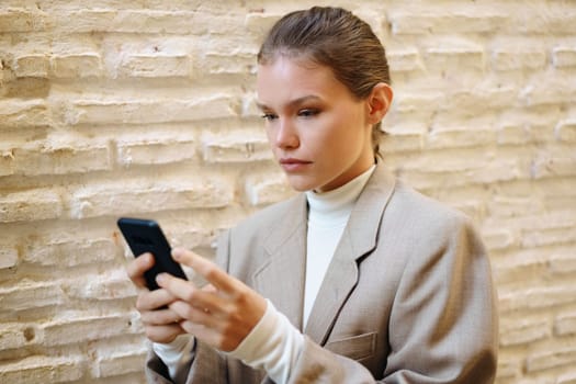 A young woman in a stylish blazer stands confidently against a textured wall, focusing on her smartphone, embodying the essence of modern lifestyle and technology in todays fastpaced world