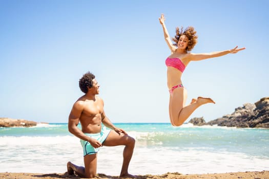 Full body of cheerful young woman in pink swimsuit jumping near kneeling African American boyfriend during vacation on beach