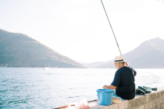 Fisherman sits on a stone fence over the sea with a fishing rod against the background of mountains. High quality photo