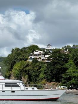 White yacht anchored near a lush tropical coastline with hillside villas and dramatic clouds overhead, evoking a serene vacation vibe.
