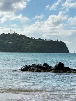 Peaceful coastal scene with dark rocks in the shallow water, clear turquoise sea, and a lush green hillside under a partly cloudy sky in the tropical Caribbean.