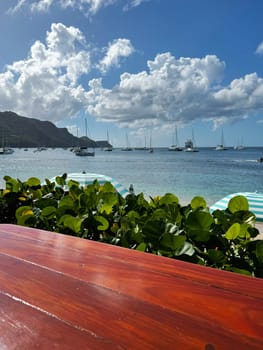 Sunny tropical coastline with sailboats in a calm bay, seen from a wooden table near beach umbrellas and lush greenery.