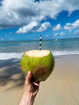 Close-up of a hand holding a green coconut with a straw on a sandy beach, with turquoise sea, blue sky, and soft white clouds in the background on a sunny day.