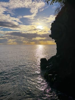 Peaceful ocean sunset with shimmering water and dramatic clouds, as a rugged rocky cliff forms a dark silhouette against the glowing horizon and colorful sky.