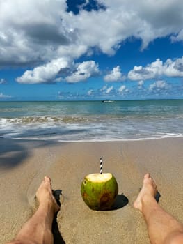 Person relaxing on sandy tropical beach with legs in the foreground, enjoying a fresh coconut drink while gazing at the calm ocean and blue sky filled with fluffy clouds.