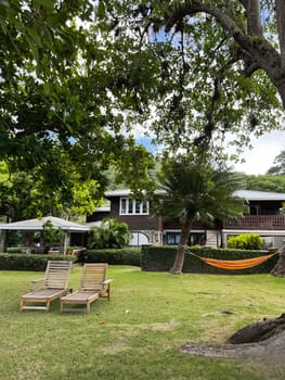 Relaxing garden scene with wooden sun loungers and a colorful hammock tied between palm trees, in front of a tropical villa surrounded by lush greenery and shade.