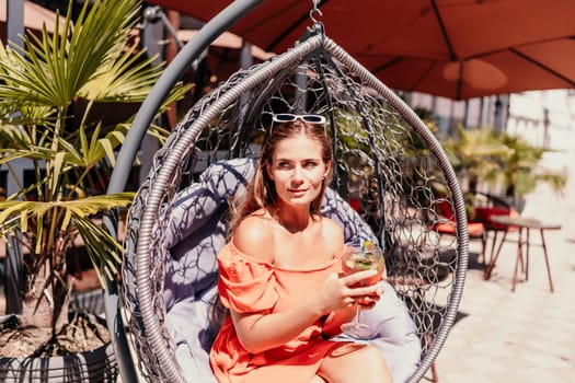 A woman in an orange dress is sitting in a hanging chair. She is holding a wine glass and smiling. The scene takes place in a restaurant with several chairs and tables