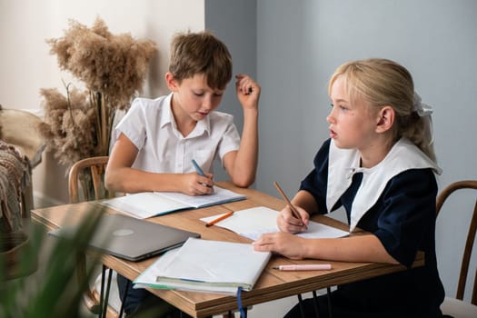 Two children, a boy and a girl, sit at a table doing homework. The boy is writing in a notebook and the girl is writing in a notebook. A laptop and a few pencils are also on the table.