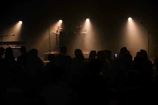Audience gathers in a lively venue, eagerly anticipating the performance as stage lights illuminate the setup, creating a vibrant atmosphere filled with excitement and energy.