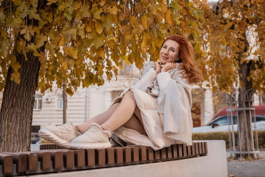 Redhead woman in a light coat sits on a park bench under autumn leaves, smiling. She wears sneakers.
