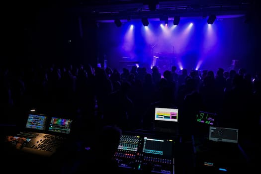 A lively concert venue filled with an energized crowd under the mesmerizing glow of blue lights. Sound equipment and vibrations fill the air as anticipation builds.