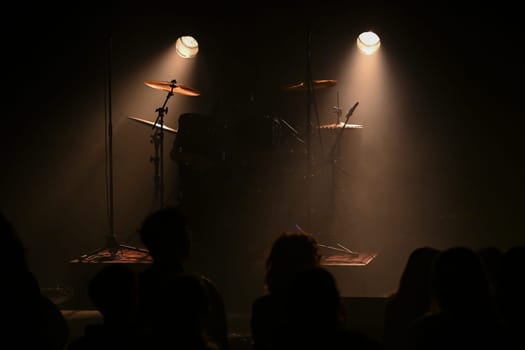 Bright stage lights illuminate the drum set as an excited crowd waits for the performance to begin, creating an atmosphere full of energy and anticipation.