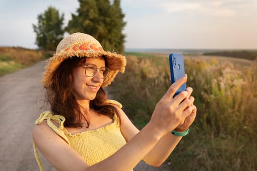 A smiling woman in a straw hat takes a selfie in a sunny rural landscape.