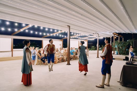 Men and women in national Montenegrin costumes dance bouncing on stage in front of guests sitting on chairs. High quality photo