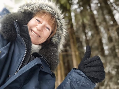 A middle-aged tourist happily poses for a selfie, giving a thumbs up while dressed warmly in a snowy forest, capturing her travel joy.