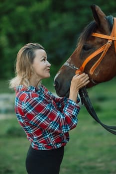 Happy blonde with horse in forest. Woman and a horse walking through the field during the day. Dressed in a plaid shirt and black leggings