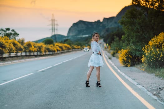 A woman in a white dress stands on a road. The road is empty and the sky is orange