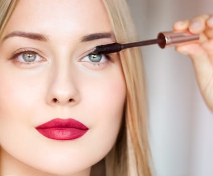 Woman applying mascara with confidence, defined brows and red lipstick. Blonde beauty focused on her eye makeup, cosmetics look