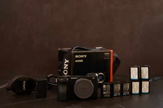 Sony A6500 mirrorless camera with charger, batteries and strap stands on a black table. High quality photo