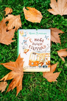 Children book with pictures lies on the lawn among fallen leaves. Title: Old women fell from the sky. Masha Rupasova. High quality photo