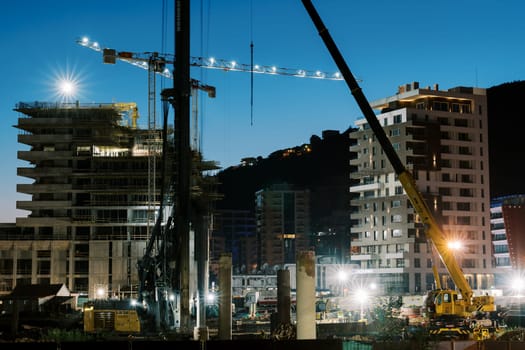 Illuminated tower cranes work at the construction site of multi-storey buildings. High quality photo