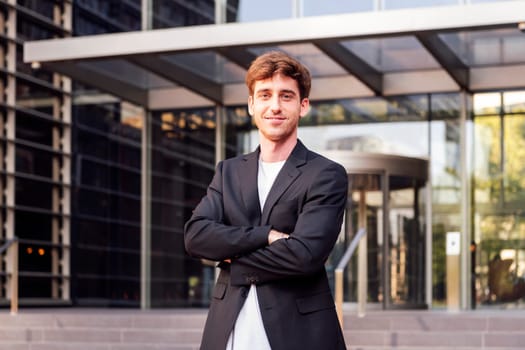 young adult man in casual suit standing looking at camera with arms crossed in front of an office building, concept of entrepreneur and business, copyspace for text