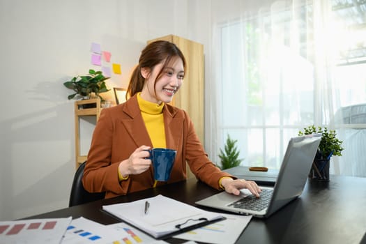 Confident young businesswoman holding a coffee mug and using laptop in a stylish home office.