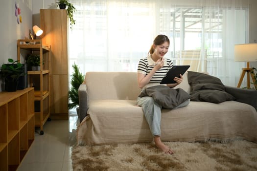 Smiling woman sitting barefoot on a cozy sofa browsing internet on digital tablet.