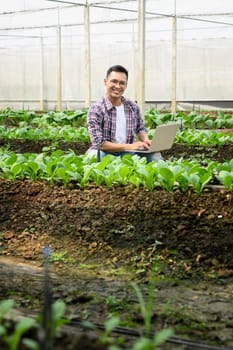 Smiling male farmer using laptop while collecting data and observing plant growth in a greenhouse.
