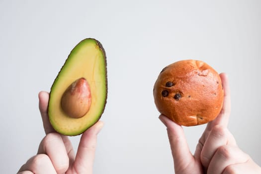 Hands holding half avocado and chocolate chip bun against white background.