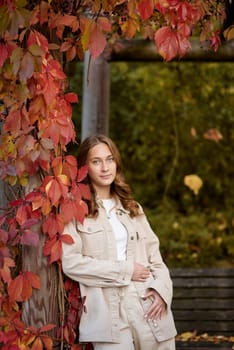 A radiant young woman in a casual beige ensemble leans gracefully against a rustic wooden pergola adorned with vibrant red autumn leaves. The warm, golden tones of fall foliage create a natural backdrop, highlighting her joyful smile and the inviting atmosphere of the garden setting. This image captures the essence of autumn lifestyle, combining modern casual fashion with the organic textures of seasonal nature. Perfect for promoting outdoor living, fashion blogs, travel content, or autumn-themed marketing campaigns, the scene evokes feelings of relaxation, connection with nature, and the comforting charm of the fall season. A young woman with wavy light brown hair and a cheerful smile is leaning against a wooden pillar wrapped in red autumn leaves. She is dressed in a beige casual outfit, including a stylish denim jacket and matching pants, with a white top underneath. The background features a scenic garden pergola with wooden beams covered in lush green and red foliage, creating a natural and cozy atmosphere. The warm lighting and soft depth of field enhance the autumn aesthetic, making this image perfect for seasonal fashion, lifestyle blogs, outdoor portraiture, and autumn-themed marketing campaigns.