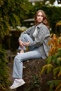 A young woman with wavy brown hair and blue eyes poses in a serene autumn park, sitting on a rustic stone wall surrounded by lush green and orange foliage. She wears a black and white checkered shirt over a white top, paired with light blue jeans and white sneakers. Her confident and relaxed expression, combined with soft natural lighting, creates a harmonious and stylish outdoor portrait. This image is ideal for autumn fashion promotions, lifestyle blogs, seasonal advertising, and social media campaigns.