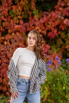 A young woman in a casual autumn outfit poses confidently against a vibrant red ivy wall, creating a stylish fashion portrait in warm seasonal tones. The cozy fall atmosphere and urban outdoor setting highlight trendy seasonal style, making this image perfect for fashion blogs, lifestyle content, seasonal marketing campaigns, or travel-related projects. The rich red tones of the ivy wall, combined with the natural light and relaxed vibe, evoke a sense of warmth, elegance, and modern urban charm.