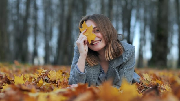 A young girl in an autumn park lies on the leaves and smiles with a leaf in her hand