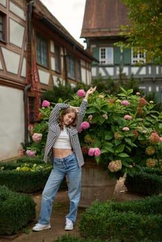 This captivating image showcases a radiant young woman in her early twenties, exuding joy and elegance in a picturesque European garden. Her casual yet trendy outfit - a white crop top layered under an unbuttoned checkered shirt, paired with relaxed blue jeans - embodies effortless autumn style. The model's playful pose, with one arm stretched upward, conveys a sense of freedom and happiness, perfectly complementing the serene countryside setting. Vibrant pink hydrangeas in a large planter add a pop of color, while the background features charming half-timbered houses with brown frames, white walls, and green shutters, epitomizing traditional European architecture. Lush autumn foliage and creeping ivy enhance the warm, inviting atmosphere, bathed in soft natural light. This versatile image is ideal for travel blogs, lifestyle magazines, fashion lookbooks, and campaigns celebrating European culture, rural charm, and the beauty of everyday moments in a picturesque village setting. Stylish Young Woman in Checkered Shirt and Crop Top Poses with Pink Hydrangeas by Half-Timbered Houses. Carefree Fashion Portrait Captures Rural Village Charm and Vibrant Fall Foliage in Picturesque Garden Setting.