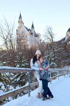 This photograph captures a beautiful woman and a child standing on a bridge in the snow, with the iconic Neuschwanstein Castle prominently featured in the background. The scene exudes a serene and picturesque winter atmosphere, with the majestic castle and the snowy landscape creating a stunning backdrop. The woman and child are dressed in warm winter clothing, adding to the cozy and inviting feel of the image. The photograph is perfect for themes related to family, winter travel, nature, and scenic landscapes, offering a detailed and evocative representation of a winter wonderland. Beautiful woman and child standing on a bridge in the snow with Neuschwanstein Castle in the background, capturing a serene winter scene with a majestic castle and snowy landscape.