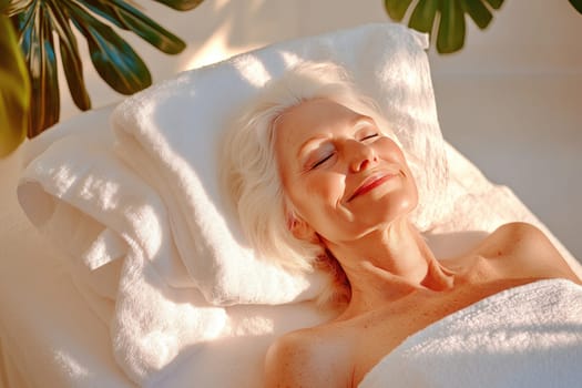 An elderly woman lies back comfortably in a calming spa setting, indulging in a facial massage. Her eyes are closed, and a serene smile adorns her face as she relaxes, with her head resting on a soft, folded towel. The clean white interior, complemented by gentle shadows, enhances the tranquil atmosphere. This peaceful moment captures the essence of self-care and rejuvenation, allowing her to unwind completely in the soothing environment.