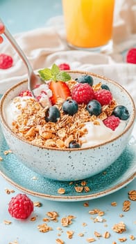 A hand reaches in with a spoon, scooping a creamy bite from a beautifully arranged granola and yogurt bowl. This bowl, resting on a rustic ceramic plate with charming glaze imperfections, is adorned with scattered granola, crushed nuts, and vibrant fresh berries. Soft refractions from a glass of orange juice add depth to the scene, enhanced by natural lighting that highlights the yogurt and glossy fruits. A blurred linen napkin and coffee cup complete the cozy breakfast aesthetic.