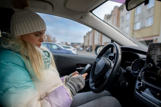 A young woman, dressed in warm and cozy attire, is actively engaging with her smartphone while sitting inside a car