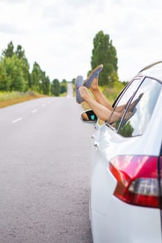 A young girl driving a car, pulling her legs out of the window, resting behind the wheel. Road trip