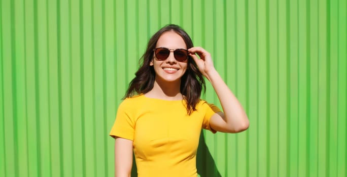 Portrait of beautiful happy smiling brunette young woman with glasses in yellow dress posing on colorful green wall background